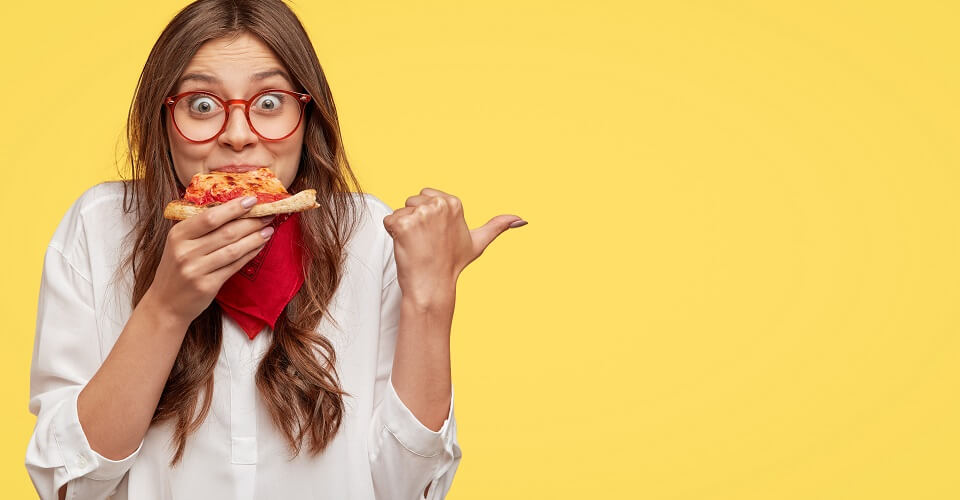 Lunch time concept. Attractive cheerful woman eats delicious Italian pizza, surprised with wonderful taste, indicates with thumb aside, shows where pizzeria situated, suggests to visit and have snack