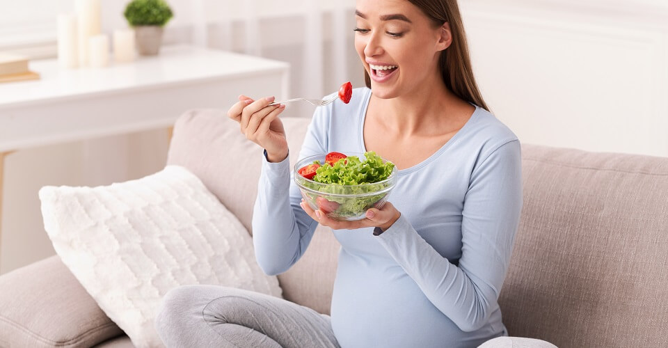 Pregnant Woman Eating Fresh Vegetable Salad From Bowl Sitting On Couch At Home. Pregnancy Nutritional Needs